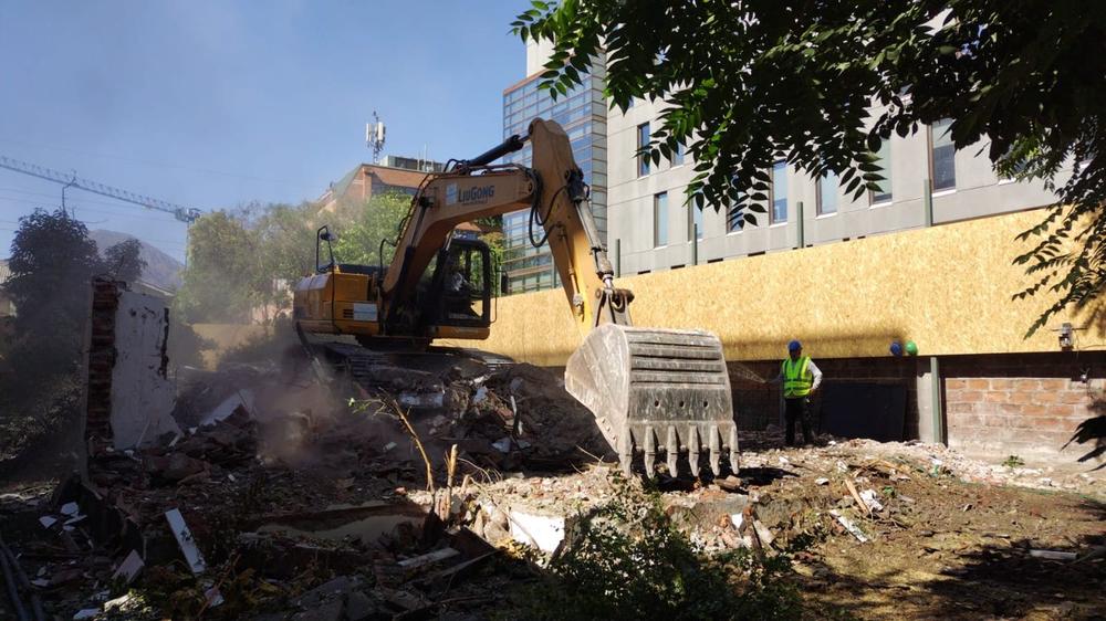 Excavadora de SG Ingeniería y Demoliciones demoliendo una vivienda junto a un edificio, con cierre perimetral de madera